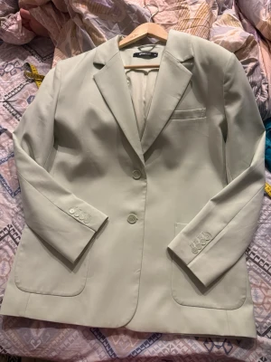 Ljusgrön kavaj från Gina Tricot - Stilren ljusgrön kavaj från Gina Tricot med klassisk passform, två knappar framtill och tre dekorativa knappar vid ärmsluten. Kavajen har två framfickor och en bröstficka, samt är helfodrad för extra komfort. Perfekt för en trendig och fräsch look.