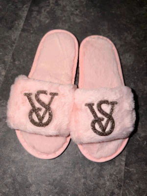 Rosa fluffiga tofflor från Victoria's Secret - Säljer ett par supermjuka och fluffiga rosa tofflor från Victoria's Secret. Tofflorna har öppen tå och bred rem med glittrigt VS-logotyp framtill. Perfekta för att chilla hemma och ge en lyxig känsla till vardagen. Storlek 36-38😍