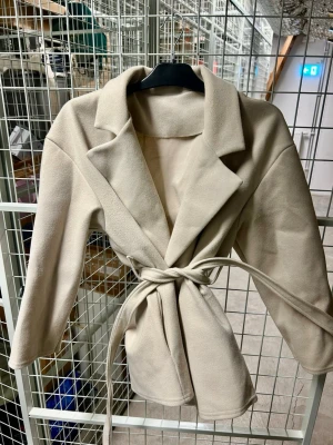 Beige kappa med knytskärp - Stilren beige kappa med bred krage och knytskärp i midjan. Kappan har en rak passform och är tillverkad i ett mjukt material som ger en lyxig känsla. Perfekt för att skapa en snygg och trendig look.