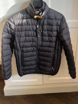 Svart dunjacka från Parajumpers - Snygg svart dunjacka från Parajumpers med quiltad design, hög krage och dragkedja framtill. Jackan har två sidofickor med dragkedja och en gul detalj vid halsen. Är en junior men passar nog en liten S eller Xs vuxen. Perfekt för våren