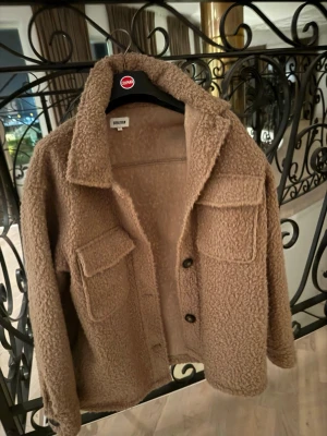 Beige teddyjacka från Gina Tricot - Mysig beige teddyjacka från Gina Tricot med två stora bröstfickor och knäppning framtill. Jackan har en avslappnad passform, klassisk krage och är gjord i fluffigt teddy-material som ger en trendig look.