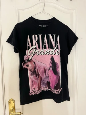 Ariana Grande Sweetener Tour T-shirt - Svart t-shirt med stort tryck av Ariana Grande och texten 'thank u, next!' på framsidan. Baksidan har turnélistan från Sweetener World Tour 2019 i rosa och vitt. Klassisk passform och korta ärmar. Perfekt för fans av Ariana Grande.