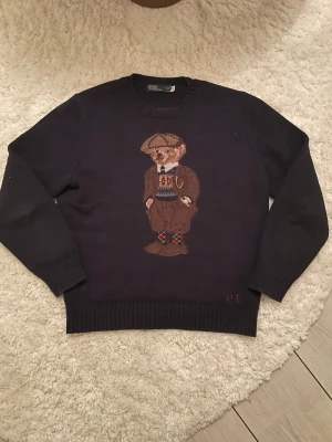 Mörkblå stickad tröja Polo Bear - Mörkblå stickad tröja från Ralph Lauren med klassiskt Polo Bear-motiv framtill. Tröjan har rund halsringning, ribbade muddar och RL-broderi vid nederkanten. Perfekt för dig som gillar preppy stil och unika detaljer.