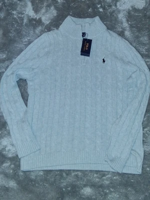  kabelstickad tröja Polo Ralph Lauren - Grå kabelstickad tröja från Polo Ralph Lauren med hög krage och ribbade muddar. Tröjan har det klassiska broderade logotypen på bröstet och är tillverkad i mjukt material. Perfekt för kyliga dagar och enkel att matcha.