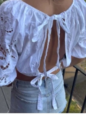 Vit blus med knyt och broderi - Supersöt vit blus med puffärmar och broderade detaljer. Toppen har öppen rygg med två knytband och är croppad i modellen. Perfekt för sommarens alla tillfällen och ger en romantisk vibe. Luftigt bomullsmaterial och fina volangdetaljer vid ärmslut och nederkant.