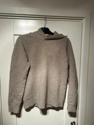 Beige hoodie i mjuk ullmix - Säljer en stilren beige hoodie i mjuk merinoull med ribbade muddar vid ärmslut och nederkant. Hoodien har en enkel design utan tryck eller fickor och en skön huva. Perfekt för dig som gillar minimalistisk stil och vill ha något bekvämt och varmt.