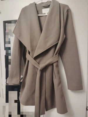 Beige kappa med knytskärp från VILA - (NY UTAN PRISLAPP)Stilren beige kappa från VILA med bred krage och knytskärp i midjan. Kappan har en rak passform, långa ärmar och är tillverkad i ett mjukt tyg som ger en lyxig känsla. Perfekt för dig som vill ha en enkel men trendig look.