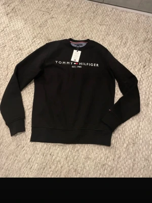Svart sweatshirt Tommy Hilfiger - Snygg svart sweatshirt från Tommy Hilfiger med klassisk logga och texten 'EST. 1985' på bröstet. Tröjan har rund halsringning, långa ärmar och ribbade muddar. Liten flaggdetalj på ärmslutet ger extra stilpoäng. PASSAR NER I STRLK