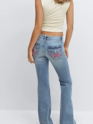 Bootcut jeans med broderade fickor - Aldrig använt med prislappen kvar på. Snygga ljusblå bootcut jeans med broderade detaljer i rosa på bakfickorna. Jeansen har låg midja och klassisk femficksdesign. Dom är försmå, det är derför jag säljer dom. 