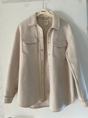 Beige kappa / overshirt från ONLY - Stilren beige overshirt från ONLY med klassisk krage, tryckknappar framtill och två bröstfickor med lock. Overshirten har långa ärmar med knapp vid ärmslut och är gjord i ett mjukt, fleece-liknande material. Perfekt för lager på lager och enkel att matcha.