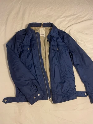 Blå jacka från J.Lindeberg - Snygg blå jacka från J.Lindeberg med beige innerfoder och detaljer. Materialet är lite glansigt och lätt, perfekt för blåsiga dagar. Storek S