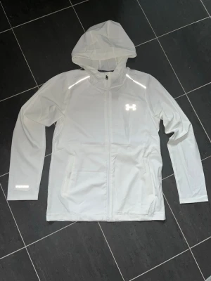 Vit vindjacka från Under Armour - Snygg vit vindjacka från Under Armour med huva och hel dragkedja framtill. Nice reflexer på den. Storlek M. Näst intill ny. Har kvar prislappen som kan följas med och till och med sättas på igen om man vill. Frågor i DM.