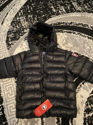 Svart dunjacka från Canada Goose - Snygg svart dunjacka från Canada Goose med huva och dragkedja. Jackan har det klassiska Canada Goose-märket på ärmen och tydlig logga på huvan. Fylld med dun för extra värme och har en modern, puffig design. Helt ny inte ens använd 
