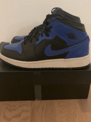 Air Jordan 1 Mid Svart/Blå hyper Royale - Säljer ett par Air Jordan 1 Mid sneakers i svart och blått läder med klassisk vit sula. Skorna har den ikoniska swoosh-loggan på sidan och Air Jordan-vingen på ankeln. Perfekta för dig som gillar streetwear och vill ha en tidlös look. Lådan tillkommer. Skick: 8/10 sparsamt använda 