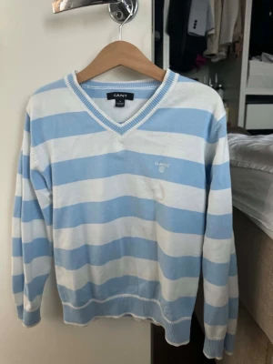 Blåvit randig stickad tröja GANT - Säljer en snygg stickad tröja från GANT med breda blå och vita ränder. Tröjan har V-ringning, långa ärmar och ribbade muddar vid ärmslut och nederkant. Perfekt för en avslappnad och stilren look.