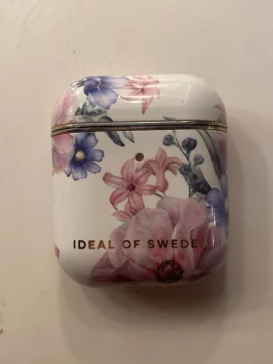 AirPods fodral Ideal of Sweden - Snyggt AirPods-fodral från Ideal of Sweden med blommigt mönster i rosa, lila och blå toner. Fodralet är gjort i hårdplast och har en blank finish samt guldfärgad text. Perfekt för att skydda dina hörlurar och ge dem en personlig touch.