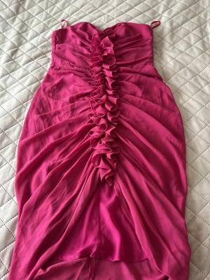 Rosa volangklänning med smala band - Supersnygg rosa klänning med smala axelband och volangdetaljer längs hela framsidan. Klänningen har en åtsittande passform och är gjord i ett tunt, lätt material som ger ett elegant fall. Perfekt för dig som vill sticka ut med färg och detaljer.