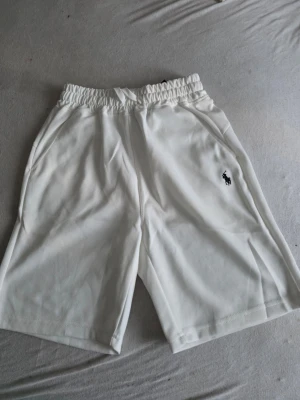 Vita shorts från Polo Ralph Lauren - Säljer ett par vita shorts från Polo Ralph Lauren med elastisk midja och sidofickor. Klassisk logga broderad i svart på ena benet. Perfekta för sommaren och har en avslappnad passform.