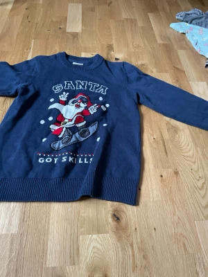 Blå jultröja med snowboardande tomte - Mörkblå stickad tröja från Kappahl med ett coolt julmotiv av en snowboardande tomte och texten 'Santa got skills'. Tröjan har ribbade muddar vid hals, ärmslut och nederkant. Perfekt för dig som vill sticka ut på julfesten!