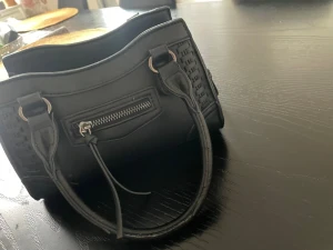 Svart handväska från Gina Tricot - Snygg svart handväska från Gina Tricot med flätade detaljer på sidorna, silverfärgade metalldetaljer och dragkedja framtill. Väskan har två handtag och flera fack inuti för smart förvaring. Perfekt för dig som gillar stilrena accessoarer.