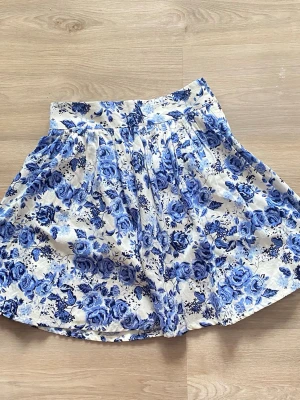 Vit kortkjol med blåa blommor - Supersöt kortkjol i vitt med blåa blommor och hög midja. Kjolen har ett utställd passform och är gjord i mjuk bomull, perfekt för varma dagar. Blommönstret ger en fräsch och somrig vibe.