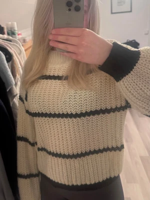 Randig stickad tröja i offwhite och svart - Säljer en chunky stickad tröja i offwhite med svarta horisontella ränder. Tröjan har en hög hals, breda ärmar och ribbade muddar vid ärmslut och nederkant. Perfekt för lager på lager och riktigt mysig till hösten och vintern.