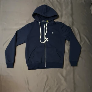 Marinblå hoodie från Polo Ralph Lauren - Snygg marinblå hoodie från Polo Ralph Lauren med dragkedja framtill och vit broderad logga på bröstet. Hoodien har vita snören i huvan och två fickor framtill. Tillverkad i mjukt bomullsmaterial som känns skönt mot huden.