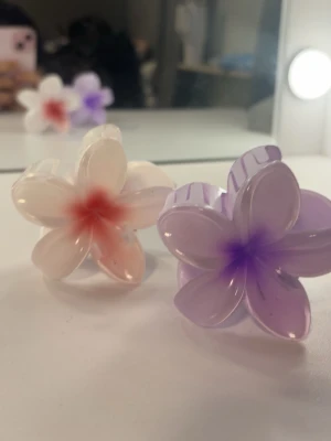 Blommiga hårklämmor i pastell - Två hårklämmor i form av blommor, en i ljusrosa med röd mitt och en i ljuslila med lila mitt. Båda är gjorda i glansig plast och har en söt, feminin look som piffar upp vilken frisyr som helst.