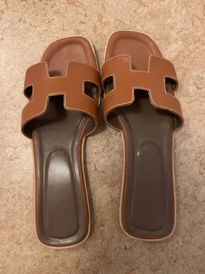 Bruna slip-in sandaler H - Snygga bruna slip-in sandaler med bred rem i H-form och vita sömmar. Sandalerna har fyrkantig tå och platt sula i brunt och beige. Tillverkade i konstläder med bekväm innersula. Perfekta för en avslappnad och trendig look.