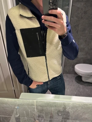 Beige fleeceväst med svart detalj - Säljer en beige fleeceväst med dragkedja och svart bröstficka med zip. Riktigt snygg nu till våren. Jag på bilden är 188cm och bär storlek S. Hör av er vid frågor och funderingar:)
