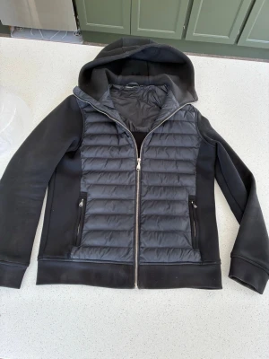 Svart cardigan - Snygg svart cardigan med huva och dragkedja framtill. Jackan har quiltad front, släta ärmar och två fickor med dragkedja. Perfekt för våren och sommar . Materialet är en mix av vadderad polyester och mjuk bomull. Otroligt snygg 