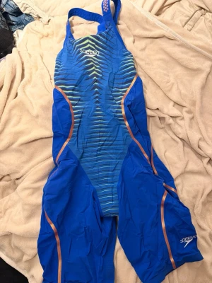 Blå tävlingsbaddräkt från Speedo - Säljer en blå tävlingsbaddräkt från Speedo med orange detaljer och randigt mönster framtill. Dräkten har breda axelband och är designad för simning på hög nivå. Materialet är snabbtorkande och tight för bästa prestanda i vattnet. Speedo tävlingsbaddräkt väl använd och har ett litet hål på magen. Dräkten går fortfarande och använda men jag valde att köpa en ny istället. Baddräkten är stl uk 28. Fina märket sitter kvar.  Pris 1000kr