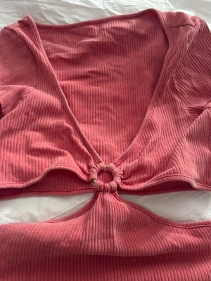 Ribbad korallfärgad croppad topp - Trendig ribbad topp i korall med korta ärmar och djup v-ringning. Toppen har en unik cut-out detalj framtill med ring och omlott-design. Perfekt för dig som vill sticka ut med en cool och modern stil. Aldrig använd, endast testad. Storlek L men passade som S/M - för liten i bysten för mig som visas i tredje bilden och jag har oftast B/C kupa