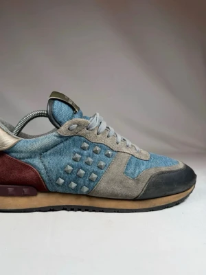 Valentino rockrunners  - Snygga sneakers från Valentino Garavani i blå denim med grå mockadetaljer och svarta partier vid tån. Skorna har coola nitar på sidorna och vinröda detaljer vid hälen. Tjock sula i beige och svart. Perfekt för dig som vill sticka ut med en unik och trendig look.