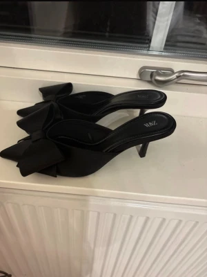 Svarta pumps med rosett från Zara - Säljer ett par svarta pumps från Zara med spetsig tå och stor dekorativ rosett framtill. Skorna har öppen häl och smal klack, vilket ger en elegant och trendig look. Materialet är slätt och matt, vilket gör dem stilrena och lätta att matcha.