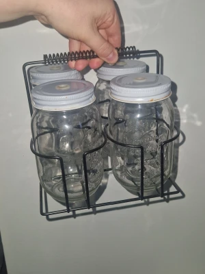 Glasmuggar m ställ  - Säljer fyra genomskinliga glasmuggqr med vita metallock, perfekta för förvaring eller dekoration. Burkarna står i ett svart metallställ med handtag, vilket gör dem enkla att bära och snygga att ha framme i köket eller på picknick. Rostade förekommer- se bilder men går att få bort med olika metoder. Kan även drickas ur utan lock. 