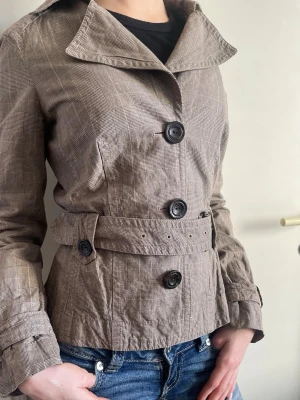 Esprit vintage vårjacka med knappar - Snygg kort gråbrun trenchcoat med bälte från Esprit. Vintage och i gott skick utan defekter!  Storlek M men passar också S och XS. Jackan har ett bälte i midjan samt ärmslejfar med spänne. Framtill finns tre stora knappar och kragen är klassisk och elegant. Perfekt jacka till våren och lätt att matcha! 