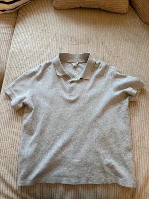 Ljusgrå piké t-shirt med krage - En stilren ljusgrå piké t-shirt med klassisk krage och två knappar framtill. T-shirten har korta ärmar och är tillverkad i mjuk bomull som känns skön mot huden. Perfekt för en clean och enkel look.