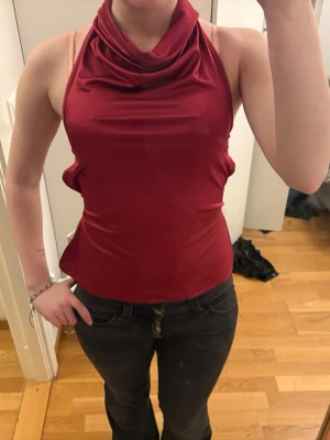 Vinröd halterneck topp med snörning - Snygg vinröd halterneck topp i glansigt material med draperad krage och öppen rygg med snörningsdetaljer. Toppen är ärmlös och har en tight passform som framhäver figuren. Perfekt för dig som vill sticka ut med en unik och trendig look.