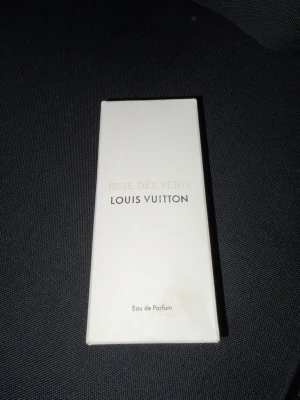 Louis Vuitton Rose Des Vents parfym - Lyxig parfym från Louis Vuitton, Rose Des Vents, i en elegant vit kartong med guld- och svart text. Förpackningen har en stilren, rektangulär form och är tillverkad av kartong. Eau de Parfum som utstrålar exklusivitet och klass.