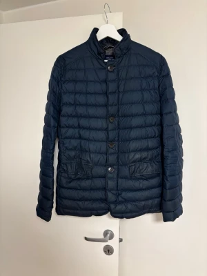 Mörkblå pufferjacka från gant perfekt nu till våren - Snygg mörkblå pufferjacka med ståkrage och knäppning framtill. Jackan har två stora fickor med lock och quiltad design som ger en modern look. Perfekt för lite kyligare dagar som nu på våren. Materialet är lätt och isolerande. Jag är 183 och den sitter bra på mig men är nyfiken på nått nytt.