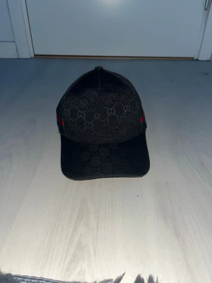 Svart Gucci keps med GG-mönster - Snygg svart keps från Gucci med klassiskt GG-monogram över hela kepsen och en grön-röd rand på sidan. Kepsen har böjd skärm och justerbar rem bak. Materialet ser ut att vara canvas med detaljer i tyg. Perfekt för dig som vill ha en exklusiv och trendig look.