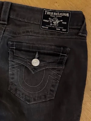 True Religion svarta bootcut jeans - Säljer ett par svarta jeans från True Religion med bootcut passform och washed look. Modellen heter Joey Low Rise Flare och har ikoniska fickor med lock och silverfärgade knappar bak. Jeansen är tillverkade i stretchigt denim.
