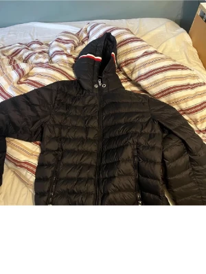 Moncler jacka  - Snygg svart dunjacka från Moncler med huva och dragkedja framtill. Jackan har två fickor med dragkedja och Moncler-logga på ärmen. Klassisk quiltad design och rödvit detalj på huvan. Perfekt för kalla vinterdagar.