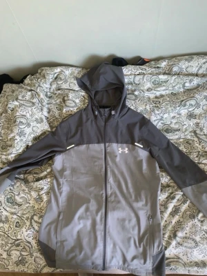 Grå vindjacka från Under Armour - Snygg grå vindjacka från Under Armour med huva och hel dragkedja framtill. Jackan har två olika grå nyanser, reflekterande detaljer och logga på bröstet samt stor logga på ryggen. Perfekt för blåsiga dagar och sportiga outfits.