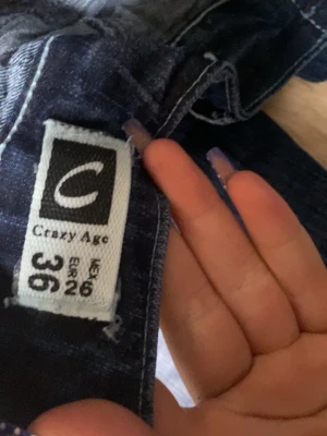 Mörkblå jeans Crazy Age strl 36 - Säljer ett par mörkblå jeans från Crazy Age i storlek 36. Jeansen har snygga slitningar, broderade detaljer och en cool logga på fickan. Klassisk femficksmodell med knapp och dragkedja. Perfekta för dig som gillar en trendig och avslappnad stil.