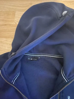 Marinblå hoodie från Sail Racing - Mörkblå hoodie från Sail Racing med dragkedja framtill och vit logga på bröstet. Tröjan har huva och ribbade muddar vid ärmslut och nederkant. Tillverkad i mjukt material som känns skönt mot huden. Perfekt för en avslappnad stil