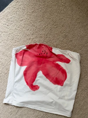 Vit tubtopp med röd blomma - Säljer en vit axelbandslös tubtopp med en stor röd blommig print framtill. Perfekt för sommardagar och enkel att styla med jeans eller kjol. Toppens material känns mjukt och stretchigt.