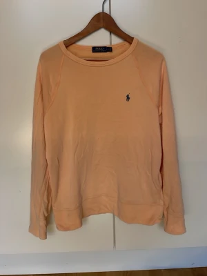 Persikofärgad sweatshirt Polo Ralph Lauren - Persikofärgad sweatshirt från Polo Ralph Lauren med klassisk rund halsringning och den ikoniska lilla loggan broderad på bröstet. Tröjan har långa ärmar och är gjord i mjukt bomullsmaterial. Perfekt för en avslappnad och stilren look.