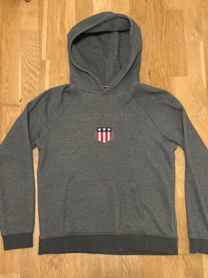 Grå hoodie från GANT med logga - Snygg grå hoodie från GANT med broderad logga och sköld med stjärnor och ränder på bröstet. Klassisk känguruficka framtill och huva. Perfekt för en avslappnad och sportig stil. Mjuk bomullsblandning som känns skön mot huden.